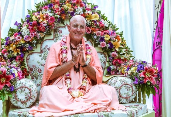 Indradyumna Swami, garlanded, seated before a flower arch